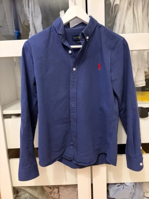 Blå skjorta från Ralph Lauren - Klassisk blå skjorta från Ralph Lauren med röd broderad logga på bröstet. Skjortan har knappar framtill och på kragen samt långa ärmar. Tillverkad i bomull och har en tidlös, normal passform.