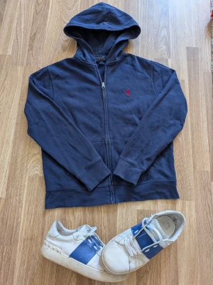 Mörkblå hoodie från Polo Ralph Lauren - Mörkblå hoodie med dragkedja från Polo Ralph Lauren. Klassisk design med huva, fickor framtill och röd broderad logga på bröstet. Tillverkad i mjukt bomullsmaterial, perfekt för en avslappnad och stilren look. Passar xs/s eller 155-165cm ca.
