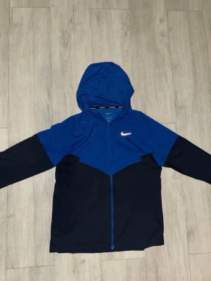 Nike Miller blå - Snygg vindjacka från Nike i blått och svart med dragkedja och huva. Jackan har en vit Nike-logga på bröstet och är tillverkad i ett lätt material som passar perfekt för blåsiga dagar. Sportig look med tvådelad färgdesign och långa ärmar.