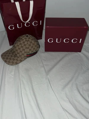 Gucci beige keps med GG-mönster - Säljer en beige keps från Gucci med det klassiska GG-monogrammet över hela kepsen. Kepsen har en böjd skärm och detaljer i grönt och rött på sidan. Tillverkad i canvasmaterial med justerbar rem bak. Perfekt för dig som vill ha en lyxig och trendig accessoar. 2025 modell