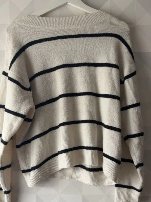 Vit stickad tröja med svarta ränder - Säljer en oversized vit stickad tröja med svarta horisontella ränder. Tröjan har rund halsringning, långa ärmar och ribbstickade muddar. Perfekt för en avslappnad och trendig look.