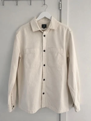 Cremefärgad overshirt/ jeansjacka - Stilren cremefärgad overshirt/ jeansjacka från H&M med två bröstfickor och svarta knappar framtill. Den har en klassisk krage och långs ärmar, perfekt för lager på lager och en clean look✨