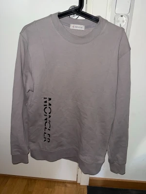 Grå långärmad tröja från Moncler - Snygg grå långärmad tröja från Moncler med svart logotyptryck längs sidan. Tröjan har rund halsringning och ribbade muddar vid ärmslut och nederkant. Perfekt för en stilren och avslappnad look.