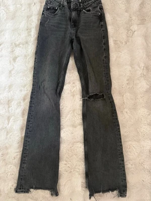 Grå raka jeans med slitningar - Säljer ett par grå raka jeans med slitna detaljer vid benslut och ett hål på ena knät. Jeansen har klassisk femficksdesign och mid waist. Perfekta för en avslappnad och trendig look.