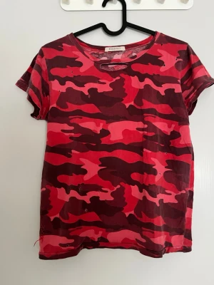 Röd camo t-shirt från RagdolL - Cool t-shirt från RagdolL med röd och rosa camouflage-mönster. Klassisk rund halsringning och korta ärmar. Perfekt för dig som vill sticka ut med en unik streetstyle-look. Tillverkad i mjuk bomull för skön känsla hela dagen.
