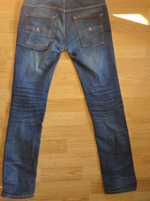 Nudie jeans - Säljer ett par mörkblå jeans med raka ben och snygga kontrastsömmar. Jeansen har två bakfickor med dekorativa sömmar och klassisk femficksdesign. Materialet är slitstark denim i bomull. Perfekta för en avslappnad och trendig look. Ett litet hål som ej syns på.