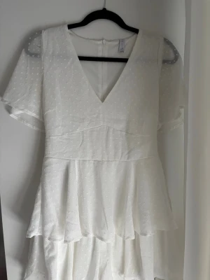 Vit prickig klänning med volanger - Söt vit klänning med små prickar, v-ringning och volangdetaljer. Klänningen har även som inbyggda  shorts (bild 2)  Klänningen har korta, transparenta ärmar. Perfekt för tex student. Eller bara på en varm sommardag klänningen har en liten fläck (bild 6) men går säkert att få bort i tvätten. 
