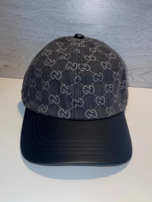  keps i denim och läder - Snygg keps i mörkgrå denim med det klassiska mönstret över hela ovandelen. Skärmen är i svart läder vilket ger en exklusiv känsla. Perfekt accessoar för dig som vill sticka ut med en lyxig och trendig look.