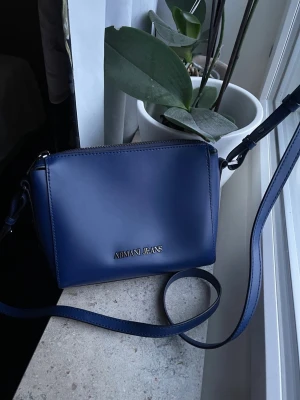 Blå handväska från Armani Jeans - Snygg blå handväska från Armani Jeans i slätt, glansigt material med justerbar axelrem. Väskan har en stilren, rektangulär form och silverfärgad logotyp framtill. Perfekt för dig som gillar enkel och modern design.helt ny