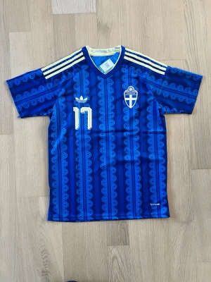 Blå Sverige fotbollströja Adidas - Säljer en blå Sverige fotbollströja från Adidas med retroinspirerat mönster, gula detaljer och nummer 17 på bröstet. Tröjan har V-ringning, broderat landslagsemblem och klassiska Adidas-ränder på axlarna. Perfekt för fotbollsfans!