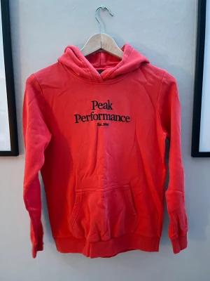 Röd hoodie från Peak Performance - Snygg röd hoodie från Peak Performance med svart logga och text på bröstet. Klassisk känguruficka framtill och skön huva. Perfekt för chill dagar eller när du vill ha en sportig look. Tillverkad i mjukt bomullsmaterial.