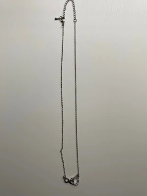 Silverfärgat halsband med hjärta och evighetssymbol - Snyggt silverfärgat halsband med en tunn kedja och ett hänge i form av ett hjärta dekorerat med små glittrande stenar och en evighetssymbol. Perfekt accessoar för att lyfta din stil med en touch av bling och symbolik.