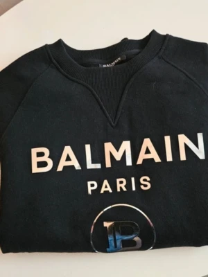 Svart sweatshirt från Balmain Paris - Som ny, använd nångång. Orginal Cool svart sweatshirt från Balmain Paris med stor vit logga framtill och rund halsringning. Tröjan har en klassisk passform och är tillverkad i mjukt bomullsmaterial. Perfekt för dig som vill ha en trendig och bekväm look med exklusiv känsla. Garanterat 100% äkta