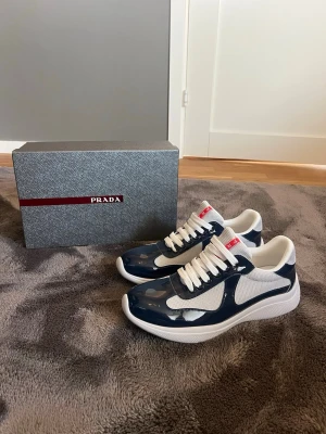 Prada cups - Snygga sneakers från Prada i marinblått och vitt med klassisk snörning. Ovandelen är i en mix av glansig syntet och mesh för en sportig look. Tydlig Prada-logga på plösen i rött. Perfekta för dig som vill ha stilrena och trendiga skor. Skriv om du har frågor :)