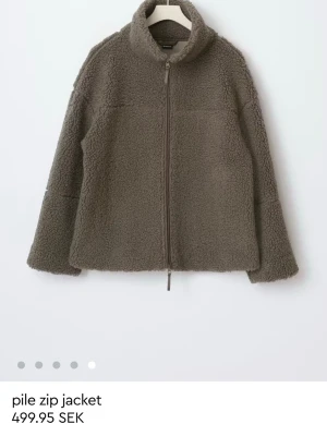 Brun teddyjacka med dragkedja - Mysig brun teddyjacka med hög krage och hel dragkedja framtill. Jackan har en avslappnad passform och är tillverkad i fluffigt pile-material som ger en trendig look. Perfekt för kyliga dagar och enkel att matcha med olika outfits.