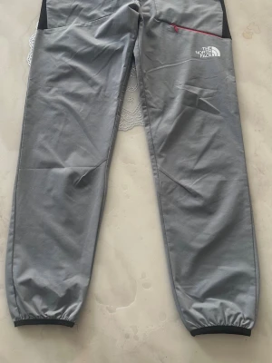 Grå mjukisbyxor från The North Face - Snygga grå mjukisbyxor från The North Face med elastiska benslut och en röd dragkedja på fickan. Loggan är tryckt på framsidan. Byxorna har en relaxed fit och är tillverkade i mjukt material som är perfekt för chill dagar.