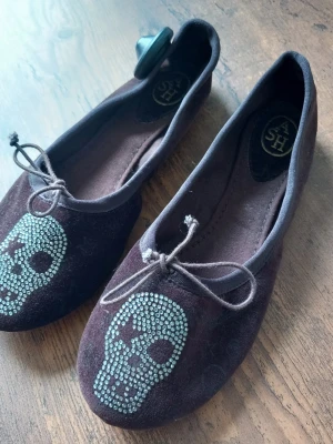 ASH Ballerinaskor Bruna med dödskalle strl 36 - Ballerinaskor med dödskallemönster i strass framtill. Skorna har en liten rosett fram och resår runt öppningen för en bekväm passform. Sulan är i gummi.