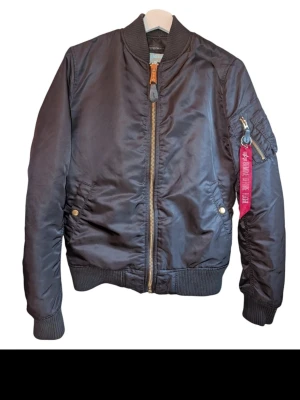 Alpha Industries Bomberjacka - Svart bomberjacka från Alpha Industries med klassiska detaljer, röd tagg på ärmen och rejäla dragkedjor.  Jackan är i använt men bra skick.  Obs! Liten i storleken!