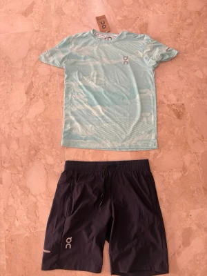 Träningsset från On – t-shirt & shorts - Snyggt träningsset från On med en ljusblå, mönstrad t-shirt i funktionsmaterial och svarta shorts med dragsko och reflexdetaljer. Perfekt för löpning eller gym. Båda plaggen har On-logga och är lätta och snabbtorkande.