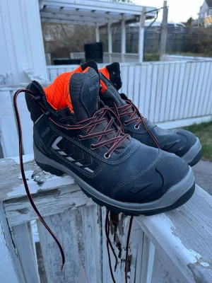 Svarta kängor från Blåkläder strl 43 - Tuffa svarta kängor från Blåkläder med orange foder och röda snören. Kängorna har robust design, förstärkta partier och detaljer i grått och vitt. Perfekta för dig som vill ha slitstarka och snygga skor med coola kontrastfärger.                            Utan låda. Orginal pris 1800kr