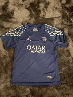PSG x Jordan blå matchtröja - Snygg blå PSG matchtröja i samarbete med Jordan. Tröjan har vita detaljer, vingar på axlarna och Qatar Airways-logga på bröstet. Tillverkad i lätt och ventilerande material, perfekt för fotboll eller träning.