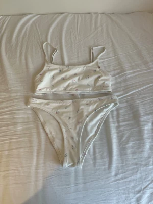 Vit bikini med småblommigt mönster - Söt vit bikini med små blommor i pastellfärger. Överdelen har smala axelband och raka kanter, medan underdelen har klassisk bikini-trosa modell. Perfekt för sommardagar på stranden eller vid poolen. En liten defekt på överdelen men inget märkbart som säkert går att få bort. 
