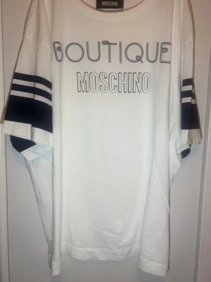 Vit oversized t-shirt från Moschino - Säljer en vit oversized t-shirt från Moschino Boutique med svart tryck framtill och breda svarta ränder på ärmarna. T-shirten har rund halsringning och en avslappnad passform som ger en cool streetwear-vibe.