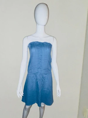 Blå sommarklänning XXL – Ny & stilren elegans - Elegant och fräsch klänning i en vacker blå nyans! 💙 Tubmodell med smock i ryggen för perfekt passform och knytdetalj i midjan. Lätt, luftigt material med diskret randig struktur – perfekt för sommar, strand eller fest. Plagget är helt oanvänt! ​Märke: Nice Fashion ​Storlek: XXL ​Mått: Bröst 54 cm, Längd 70 cm 