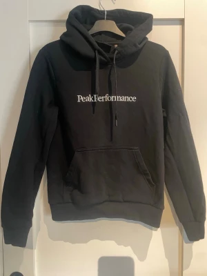 Svart hoodie från Peak Performance - Svart hoodie från Peak Performance med vit broderad logga på bröstet. Klassisk modell med huva och dragsnören samt en stor magficka framtill. Mjuk bomullsblandning och ribbade muddar vid ärmslut och nederkant. Perfekt för en avslappnad och stilren look.