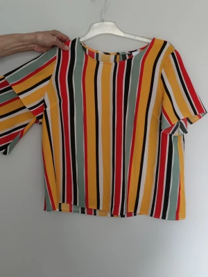 Flerfärgad randig blus - Färgglad blus med vertikala ränder i gult, rött, svart, vitt och grönt. Blusen har rund halsringning och korta ärmar. Blusen är helt oanvänd. 