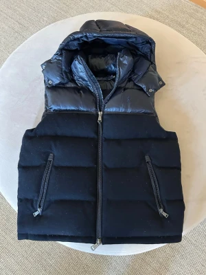 Mörkblå dun-/hybridväst från Polo Ralph Lauren - Exklusiv mörkblå dunväst från Polo Ralph Lauren med glansig ovandel och vadderad och avtagbar huva. Västen har två dragkedjefickor framtill och quiltad design. Perfekt för lager-på-lager och streetstyle. Nypris: ca 4000 kr. Mitt pris: 1750 kr