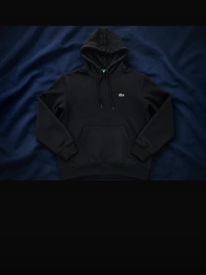 Lacoste Hoodie Black Classic Fit - Svart Lacoste hoodie i Classic Fit med broderad krokodil-logo på bröstet. Stilren och enkel hoodie som passar till allt. Bekväm passform och perfekt för både vardag och streetwear-fit.  • Storlek: M • Färg: Svart • Skick: Bra skick • Äkta Lacoste • Broderad logo • Hoodie med kangaroo pocket