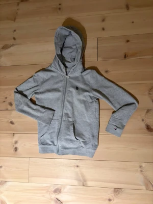 Grå hoodie med dragkedja - Snygg grå hoodie från U.S Polo Assn med dragkedja framtill, två fickor och klassisk huva. Liten broderad logga på bröstet. Perfekt för en avslappnad och stilren look.