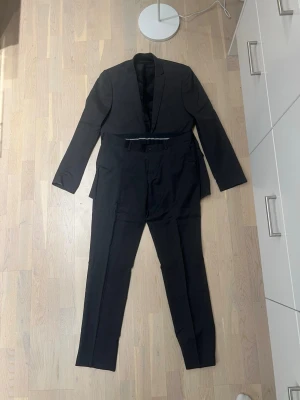Svart kostym med klassisk kavaj - Snygg svart kostym med klassisk kavaj och matchande byxor. Kavajen har smal krage och två knappar framtill. Byxorna har bälteshällor och knappstängning. Perfekt för dig som vill ha en stilren och tidlös look.