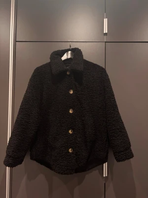 Svart teddyjacka med knappar - Mysig svart teddyjacka med stora knappar framtill och klassisk krage. Jackan har en oversized passform och är gjord i fluffigt, mjukt material. Perfekt för dig som vill ha en trendig och bekväm look.