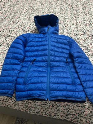 Blå dunjacka med huva från McKinley - Snygg blå dunjacka från McKinley med huva och dragkedja framtill. Jackan har två sidofickor med dragkedja och quiltad design. Perfekt för kalla vinterdagar och riktigt lätt att bära. Klassisk passform och sportig look.