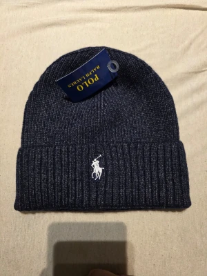 Mörkblå ribbstickad mössa Polo Ralph Lauren - Säljer en mörkblå ribbstickad mössa från Polo Ralph Lauren med klassisk vit broderad logga framtill. Mössan har uppvikt kant och är tillverkad i mjukt material som håller dig varm. Perfekt accessoar för kalla dagar.