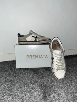 Vita Premiata sneakers med svart detalj - Snygga vita sneakers från Premiata med svarta detaljer och logga på sidan. Skorna har klassisk låg profil, vita snören och en slitstark gummisula. Tillverkade i läder och mocka för en stilren och trendig look.