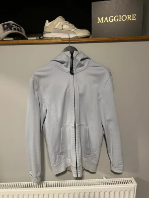 Ljusgrå hoodie med glasögon i huvan - Säljer en ljusgrå hoodie från C.P. Company med dragkedja framtill och deras ikoniska glasögon i huvan. Hoodien har två fickor på sidorna och är tillverkad i mjukt bomullsmaterial. Perfekt för dig som vill sticka ut med en unik detalj.