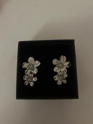 Silvriga blomformade örhängen - Unika örhängen i silverfärg med tre detaljerade blommor på varje örhänge. Blommorna har en elegant och glansig finish som ger ett exklusivt intryck. Perfekta för dig som vill ha något som sticker ut och kompletterar din stil.