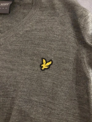 Grå stickad tröja från Lyle & Scott - Snygg grå stickad tröja från Lyle & Scott med klassisk gul fågel-logga på bröstet. Tröjan har v-ringning och är tillverkad i mjukt stickat material, perfekt för lager-på-lager-stil.