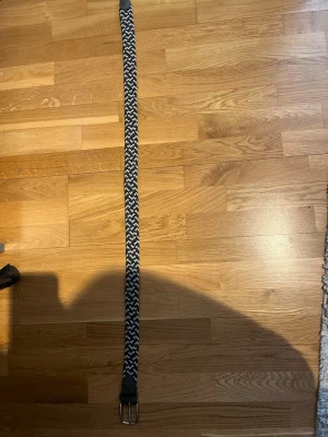 Svartvitt flätat bälte - Snyggt svartvitt flätat bälte med silverfärgat spänne. Bältet har ett randigt mönster och är gjort i ett mjukt syntetmaterial som gör det flexibelt och bekvämt att bära. Perfekt för att ge din outfit en cool touch.