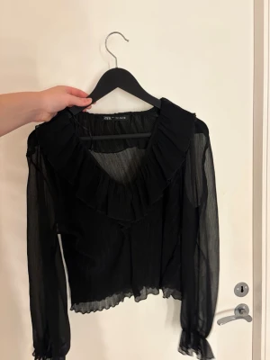 Svart transparent blus från Zara - Snygg svart blus från Zara i transparent, lätt material med volangdetalj längs den djupa v-ringningen. Blusen har långa ärmar med resår och volang vid ärmslut samt vågig kant nertill. Perfekt för dig som gillar trendiga och feminina plagg.