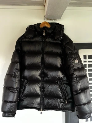 Svart dunjacka från Moncler - Säljer en svart, glansig dunjacka från Moncler med huva och två dragkedjeförsedda fickor. Jackan har en puffig design och Moncler-logga på ärmen. Perfekt för kalla vinterdagar och riktigt snygg streetstil. NFC Funkar.
