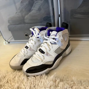 Jordan sneakers vit/lila/svart - Snygga Jordan sneakers i vitt, lila och svart med högt skaft. Skorna har detaljer i mocka och läder, samt en cool svart lackpanel på sidan. Insidan är fodrad med lila tyg och de har klassisk snörning. Perfekta för dig som gillar streetwear och basketstil.