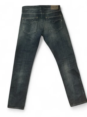 Replay blå slitna jeans herr - Snygga blå jeans från Replay med slitningar och tvättad look. Klassisk femficksmodell med raka ben och detaljerade bakfickor. Jeansen har en cool, använd känsla och Replay-lapp bak i midjan. Perfekta för en avslappnad streetstil.