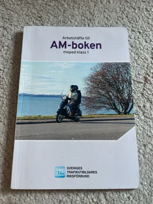 Arbetshäfte till AM-boken moped klass 1 - Arbetshäfte till AM-boken för moped klass 1, utgiven av STR. Boken har ett vitt omslag med ett foto av en person på moped vid vattnet och tydlig lila text. Perfekt för dig som pluggar till AM-körkort och vill ha kontrollfrågor och övningsmaterial.