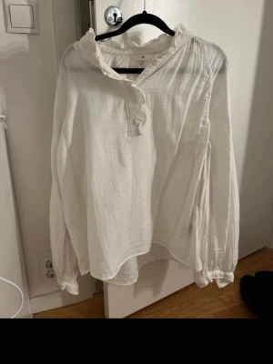 Vit blus med volang från H&M - Söt vit blus från H&M med volangkant vid halsen och knappar framtill. Tillverkad i ett lätt, luftigt tyg som ger en avslappnad känsla. Blusen har långa ärmar med resår vid ärmslut och en lös passform som passar perfekt till jeans eller kjol.