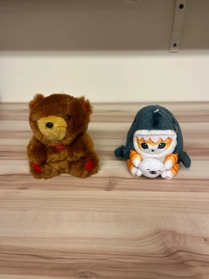Två mjukisdjur: björn & katt i hajdräkt - Säljer två mjukisdjur, en brun nallebjörn med röda detaljer och en orangevit katt utklädd i en grå hajdräkt. Båda är små, mjuka och perfekta som dekoration eller present. Materialet är plysch och de är fyllda med mjuk stoppning. Helt nya utan prislapp!