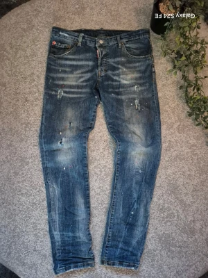 Blå dsquared2 slitna jeans med färgstänk - Säljer ett par blå jeans med distressed detaljer och färgstänk för en cool, avslappnad look. Jeansen har flera slitningar och hål på framsidan samt en tvättad finish. Perfekta för dig som gillar streetwear och en trendig stil.